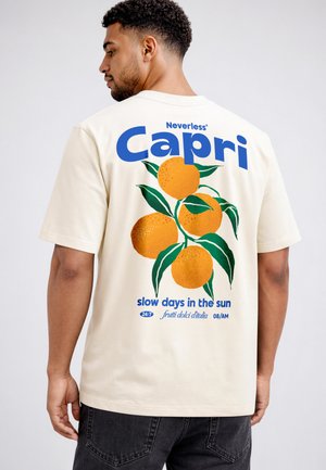 Man with short curly hair wearing cream T-shirt with large orange fruit and green leaves design and blue text "Capri" on the back.
