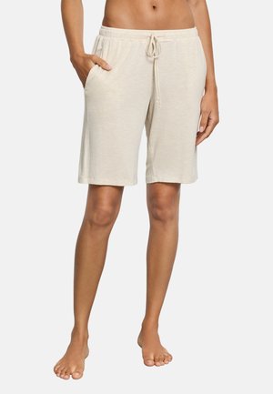 Barfodet person iført beige knælange shorts med snøre og hænderne i lommerne, stående mod en ensartet lys baggrund.