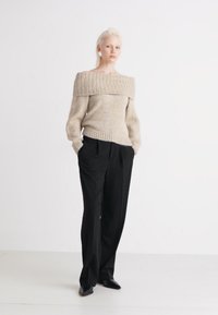 Beige oversized off-shoulder tröja med texturerad stickning och ribbstickade ärmslut, tillsammans med hög midja svarta byxor och svarta skor.