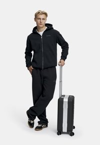 Svart zip-up hettegenser og joggebukser med logo-detaljer. Elegant svart koffert med sølvfargede detaljer og hjul, plassert ved siden av modellen.