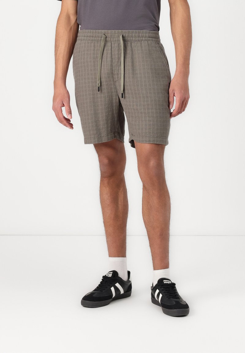 Pantalones cortos verde oliva cortos con un patrón de cuadrícula texturizado, cinturilla elástica y cordón. Combinados con zapatillas negras y blancas.