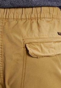 Khakihosen aus einem strukturierten Stoff, mit elastischem Bund und einer seitlichen Klapptasche, die kontrastierende Nähte zeigt.