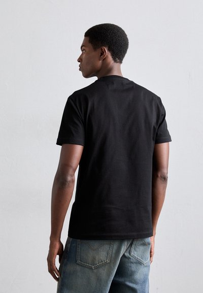 Junger Mann mit kurzen lockigen Haaren, der ein schlichtes schwarzes T-Shirt und eine blaue Jeans trägt, leicht von der Kamera abgewandt vor einem schlichten hellen Hintergrund.