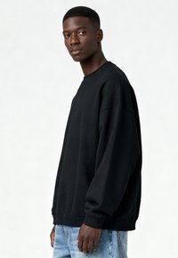 Felpa nera oversize in tessuto morbido e spesso. Presenta polsini e orlo a coste, con scollo rotondo. Indossata con jeans azzurri.