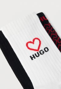 Κάλτσες από βαμβακερό μείγμα, με λευκή ριμπ κορυφή που φέρει το κείμενο "HUGO" και μια κόκκινη καρδιά, σε συνδυασμό με μαύρη βάση με σχέδιο καρδιών.
