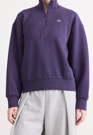 Person trägt ein violettes Sweatshirt mit Viertel-Reißverschluss und kleinem grünem Krokodil-Logo sowie locker sitzende hellgraue Hosen.