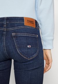 Jeans de mezclilla azul oscuro con un parche de cuero marrón marcado "TOMMY JEANS" en la cintura, que cuenta con un pequeño logotipo de color en el bolsillo trasero.