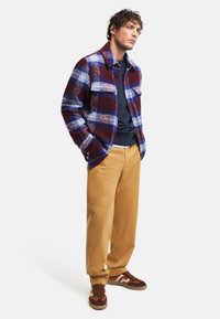 Chaqueta de cuadros en burdeos, azul y blanco, combinada con una camisa azul marino, pantalones beige y zapatillas marrones con acentos texturizados.