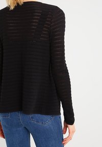 Top nero a maniche lunghe in maglia con motivo a strisce lavorate, vestibilità comoda e scollo rotondo sul retro, abbinato a jeans di denim blu.