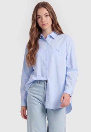 Junge Frau mit langen welligen Haaren, die ein hellblaues Hemd mit Knöpfen und hellblaue Jeans trägt, steht vor einem schlichten Hintergrund.