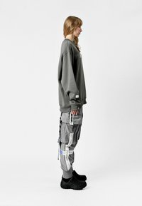 Sudadera gris de gran tamaño con puños acanalados, combinada con pantalones cargo grises que presentan múltiples bolsillos y correas con estampado. Calzado negro.
