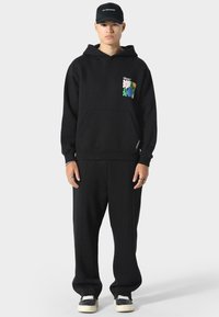 Musta huppari, jossa on etutasku, ja siinä on värikäs graafinen suunnittelu rinnassa. Yhdistetty vapaisiin mustiin verkkareihin ja lenkkareihin.