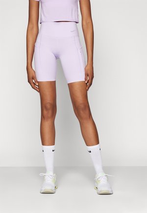 Personne portant un cuissard cycliste taille haute violet clair avec poches latérales, un crop top assorti, des chaussettes montantes blanches Nike et des baskets blanches.
