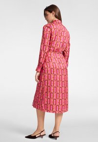 Largo vestido rosa con un patrón geométrico y acentos en naranja y púrpura. Hecho de una tela suave, con cuello y mangas largas.