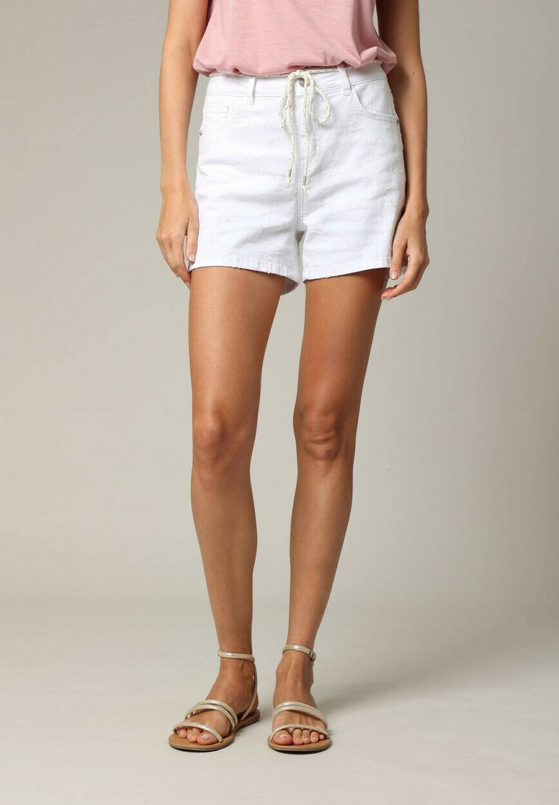 Shorts en denim blanc avec une taille à cordon, présentant un motif texturé. Associés à des sandales plates métalliques. Poches verticales visibles.