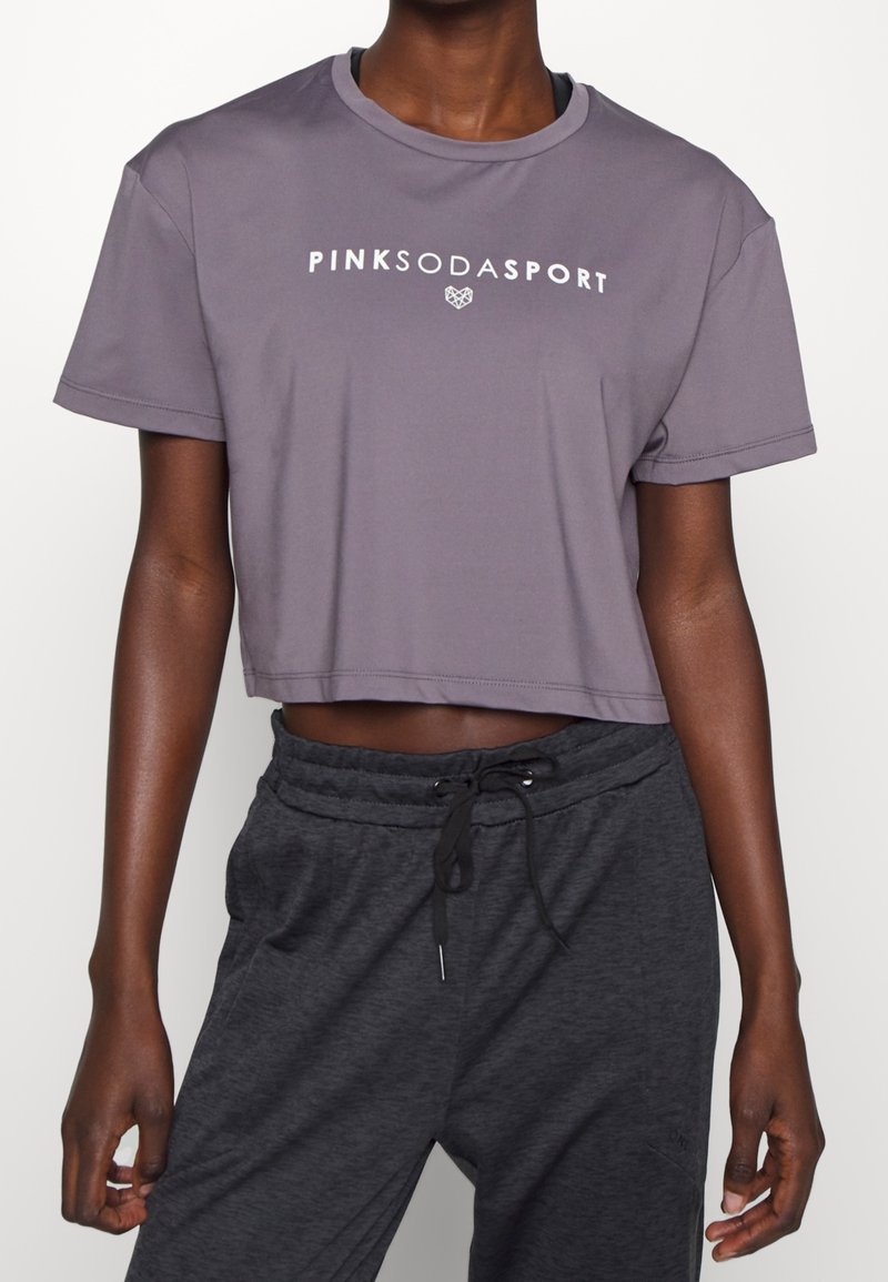Person som bär en lila kort T-shirt med texten "PINK SODA SPORT" och mörkgrå mjukisbyxor med dragsko mot en enkel vit bakgrund.