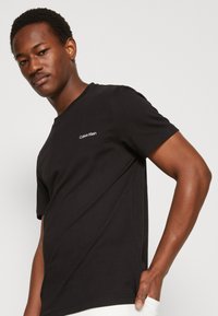 Sort bomulds T-shirt med korte ærmer, med et hvidt broderet logo på venstre bryst og med rund halsåbning.
