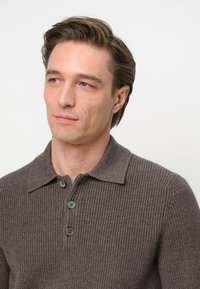 Polo de punto marrón con tejido texturizado, que presenta tres botones grises en la tapeta y un cuello. Fondo liso, enfoque en los detalles de la camisa.