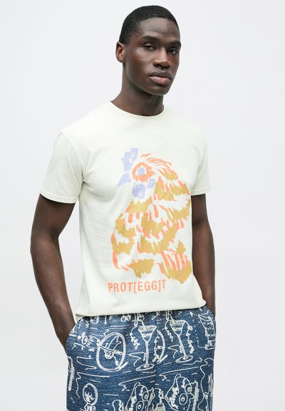 Ανοιχτοπράσινο βαμβακερό t-shirt με γραφικό σχέδιο πουλιού σε πορτοκαλί και χρυσό, σε συνδυασμό με σκούρο μπλε σορτς με σχέδια.