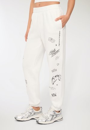 Pantaloni della tuta bianchi con vita e polsini elasticizzati, caratterizzati da diverse stampe grafiche nere, tra cui testi, ali e design di dadi.