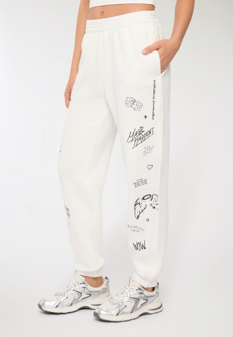 Białe dresy z elastycznym paskiem w talii i mankietami, z różnymi czarnymi grafikami, w tym tekstami, skrzydłami i wzorami kostek.