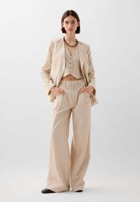 Beige gestreept pak met een cropped blazer en hoge wijde broekspijpen, met drie knopen op de gilet en twee zijzakken.