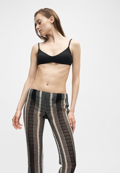 Νεαρή γυναίκα που φοράει μαύρο bralette και ψηλόμεσο πλεκτό παντελόνι με κάθετες ρίγες, πόζαρε γυρνώντας το κεφάλι της στο πλάι σε απλό φόντο.