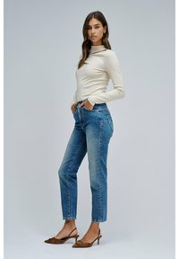 Mujer joven de pie con las manos en los bolsillos, vistiendo una blusa de manga larga color crema, jeans de tiro alto y zapatos de tacón marrones tipo slingback.