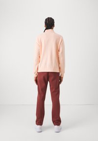 Sudadera de color rosa claro con un cuello alto, combinada con pantalones de chándal rojo oscuro. Zapatillas blancas visibles. Textura suave, diseño de ajuste relajado.