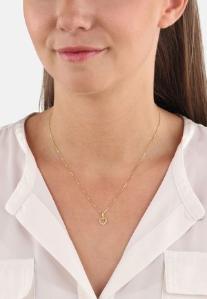 Collier en chaîne dorée avec un pendentif en forme de cœur orné de petites pierres claires, présenté sur une blouse blanche.