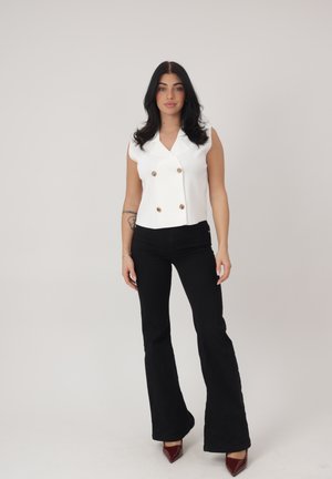 Kvinde med langt sort hår iført hvid ærmeløs, dobbeltradet vest, sorte flarebukser og bordeauxfarvede spidse hæle, stående mod en ensfarvet baggrund.