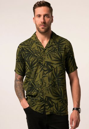 Uomo che indossa una camicia verde a maniche corte con motivo di foglie nere, pantaloni neri, orologio da polso e tatuaggio sull'avambraccio sinistro, in piedi con una mano in tasca.