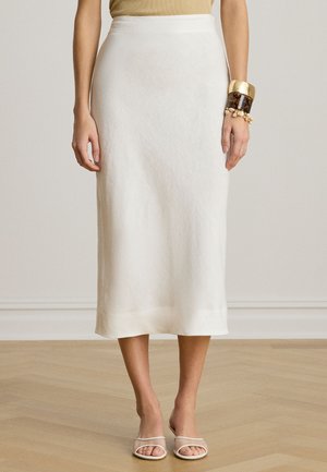 Femeie purtând o fustă midi albă, un top bej, sandale cu degetele descoperite și o brățară masivă aurie și cu model țestoasă la încheietura mâinii drepte, stând pe un podea de lemn.