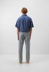 Camisa de mezclilla de manga corta, azul, con un corte relajado. Pantalones de trabajo grises a rayas con bolsillos laterales y calzado marrón claro. Fondo liso.