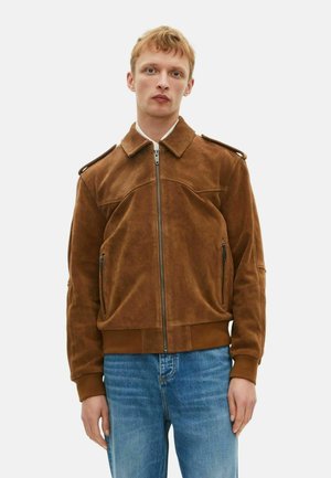 Giacca in suede marrone con zip frontale, colletto classico, spalline e tasche laterali con zip. Texture morbida, abbinata a jeans azzurri chiari.