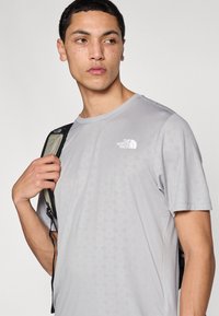 Αθλητική κοντομάνικη μπλούζα ανοιχτού γκρι με γεωμετρικό σχέδιο, κοντά μανίκια και μικρό λευκό λογότυπο στο στήθος. Το ύφασμα φαίνεται λείο και διαπνεόμενο.