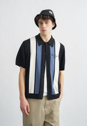 Ung mand iført sort bøllehat, sort kortærmet skjorte med blå og hvide lodrette striber og beige bukser, stående mod en hvid baggrund.