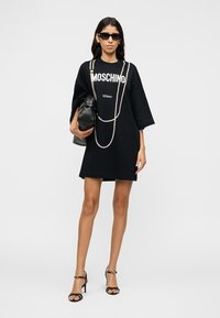 Femme portant une robe noire Moschino, des lunettes de soleil, un collier de perles, tenant un sac à main noir et des sandales à talons noirs sur un fond uni.