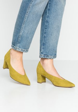 Underbenene iført lyseblå denimjeans og sennepsgule ruskind block heel pumps på et hvidt gulv.