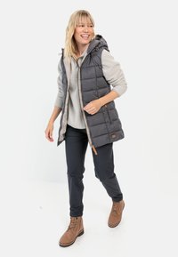 Grå polstret vest med hætte, parret med en grå fleece-top. Mørke bukser og brune snørebåndsstøvler fuldender outfitet. Lynlåsdetaljer er synlige.