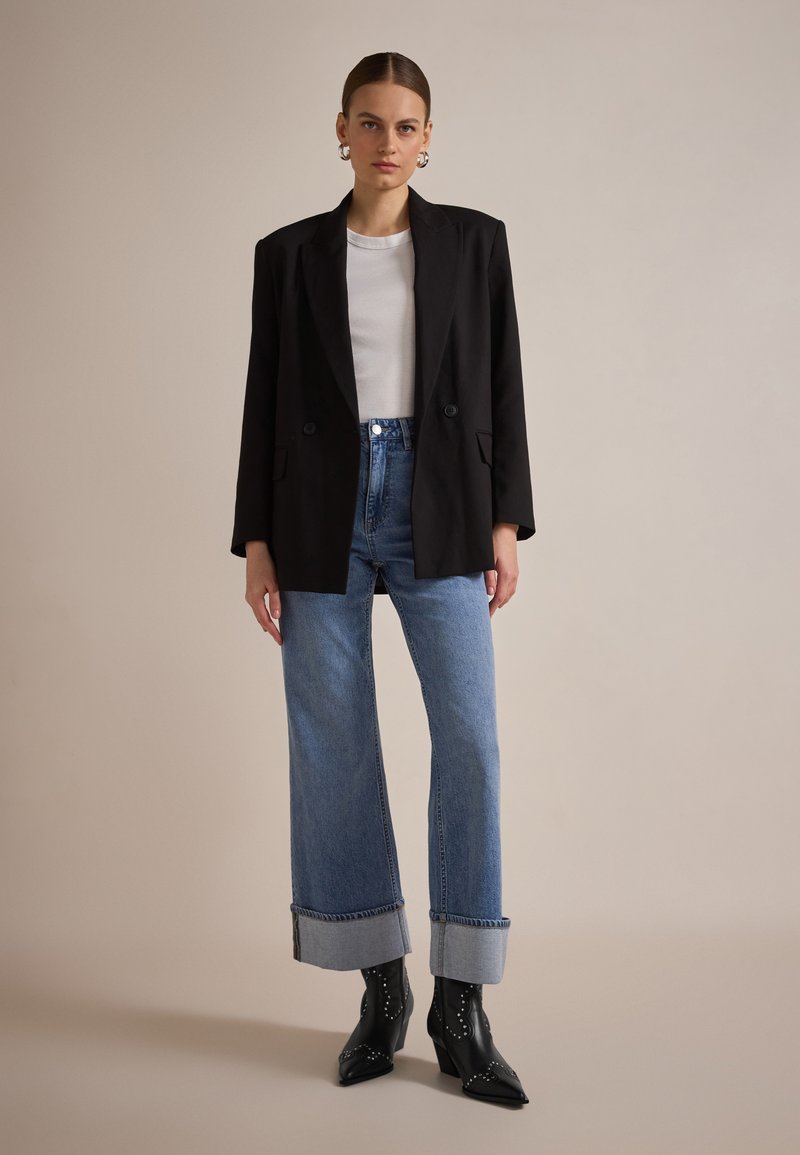 Blazer negro con dos botones, camisa blanca ajustada y jeans de cintura alta en azul con puños enrollados. Botas de tacón negro con punta afilada y tachuelas.