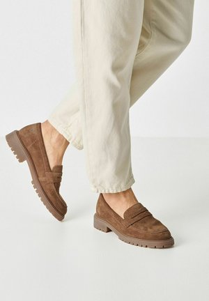 Braune Wildleder-Slipper mit klobiger Sohle, flachem Absatz und doppelter horizontaler Naht über dem Vorderblatt. Kombiniert mit hellen Hosen.