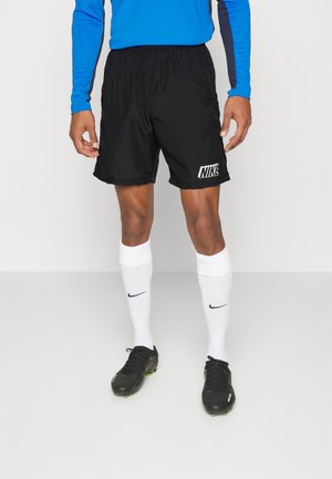 Man draagt zwarte Nike sportshorts, witte Nike kniehoge sokken, zwarte voetbalschoenen en een blauw shirt met lange mouwen.