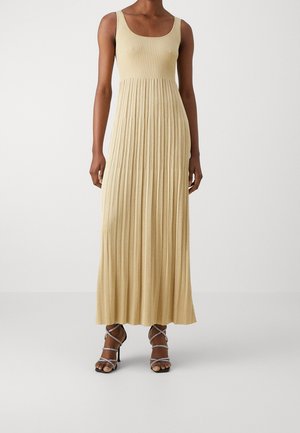 Donna che indossa un abito lungo beige senza maniche, a coste e plissettato, con sandali argentati con tacco alto e cinturini su sfondo neutro.
