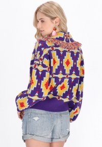 Gebreide trui met een meerkleurig geometrisch patroon in paars, geel, rood en blauw, met een franjeskraag en een ribbelrand.