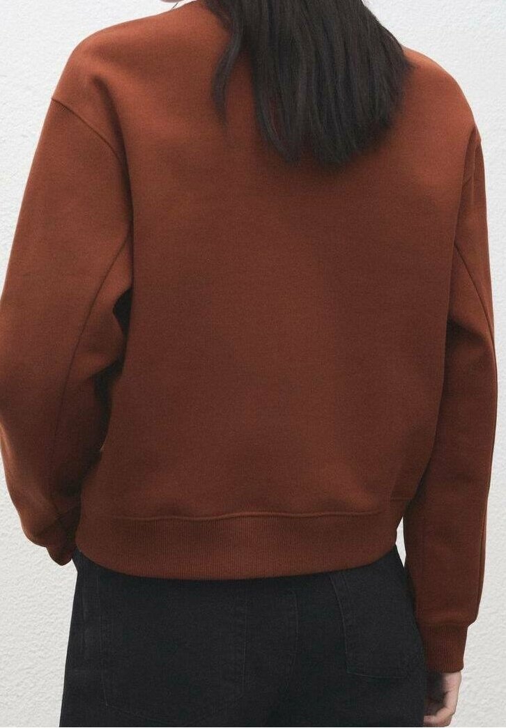Personne aux longs cheveux foncés portant un sweat-shirt couleur rouille et un pantalon foncé, debout contre un mur clair, vue de dos.