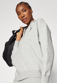 Grå zip-up sweatshirt i bomullsmaterial, med huva och känguruficka. Modellen bär en svart väska med texturerade accenter.
