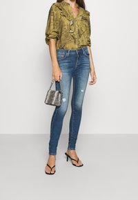 Blusa verde scuro a fantasia con scollatura arricciata, jeans skinny blu e una piccola borsa a tracolla argentata. La modella indossa sandali neri con tacco.