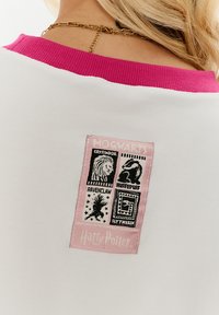Biała koszulka z różowym ribbym przy kołnierzy. Posiada prostokątną naszywkę z czarnymi symbolami domów z Hogwartsu oraz napisem "Harry Potter" w kolorze różowym.