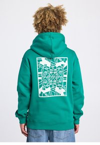 Sweat à capuche vert en coton avec une poche kangourou à l'avant. Il présente un grand motif géométrique blanc en forme de visage dans le dos.